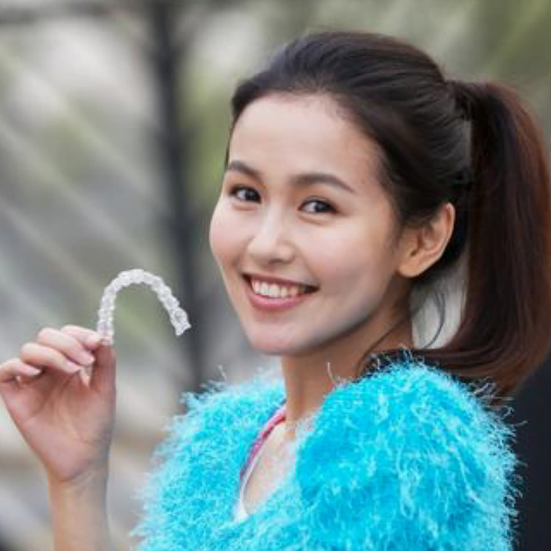 A smiling woman holds a clear dental aligner, showcasing the work of her skilled orthodontist. She's wearing a fuzzy blue top with a background of blurred plants.