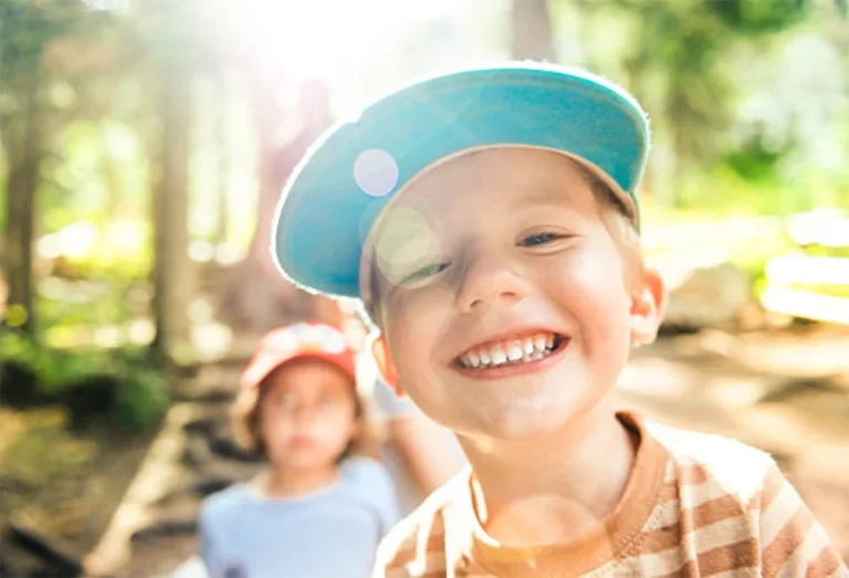 Smiling young boy in a blue cap outdoors, with another child blurred in the background on a sunny day.