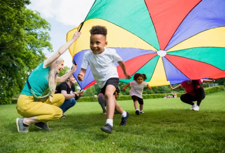 Children and adults play with a colorful parachute on a grassy field in a park.