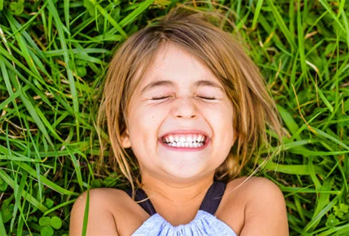 Smiling child with closed eyes lying on green grass, showing teeth and appearing joyful.