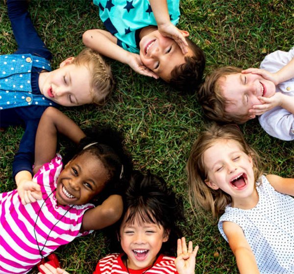 Six smiling children lie on grass in a circle, heads together, laughing and looking up at the camera.