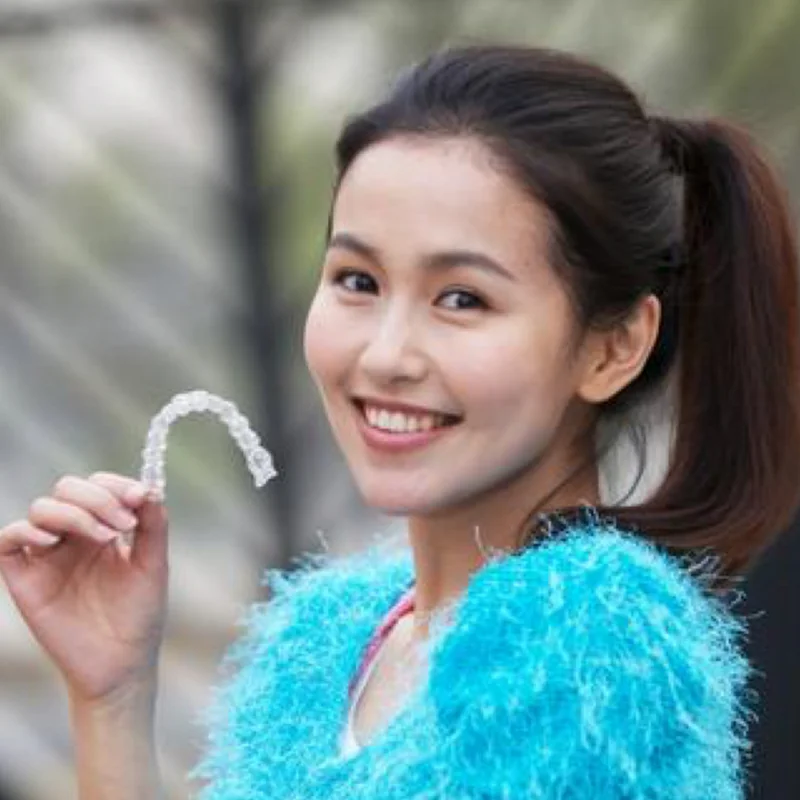 Woman with a ponytail smiling and holding a clear dental aligner, wearing a fluffy blue top outdoors.