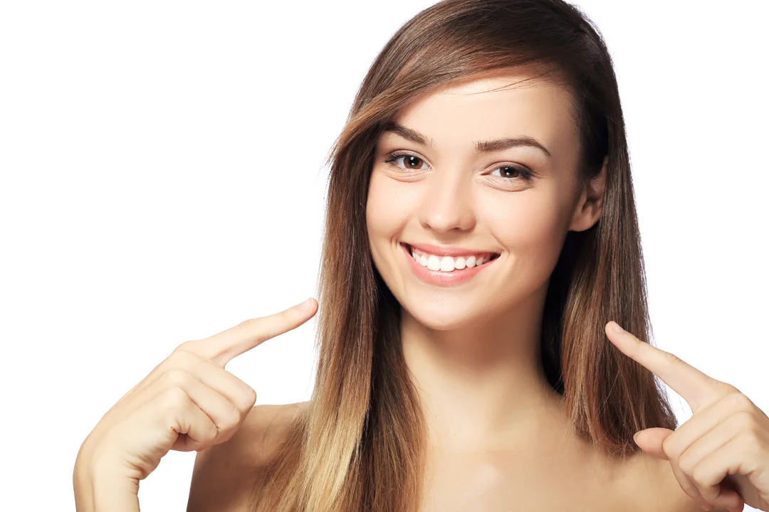 Smiling woman with long hair pointing at her teeth against a white background.