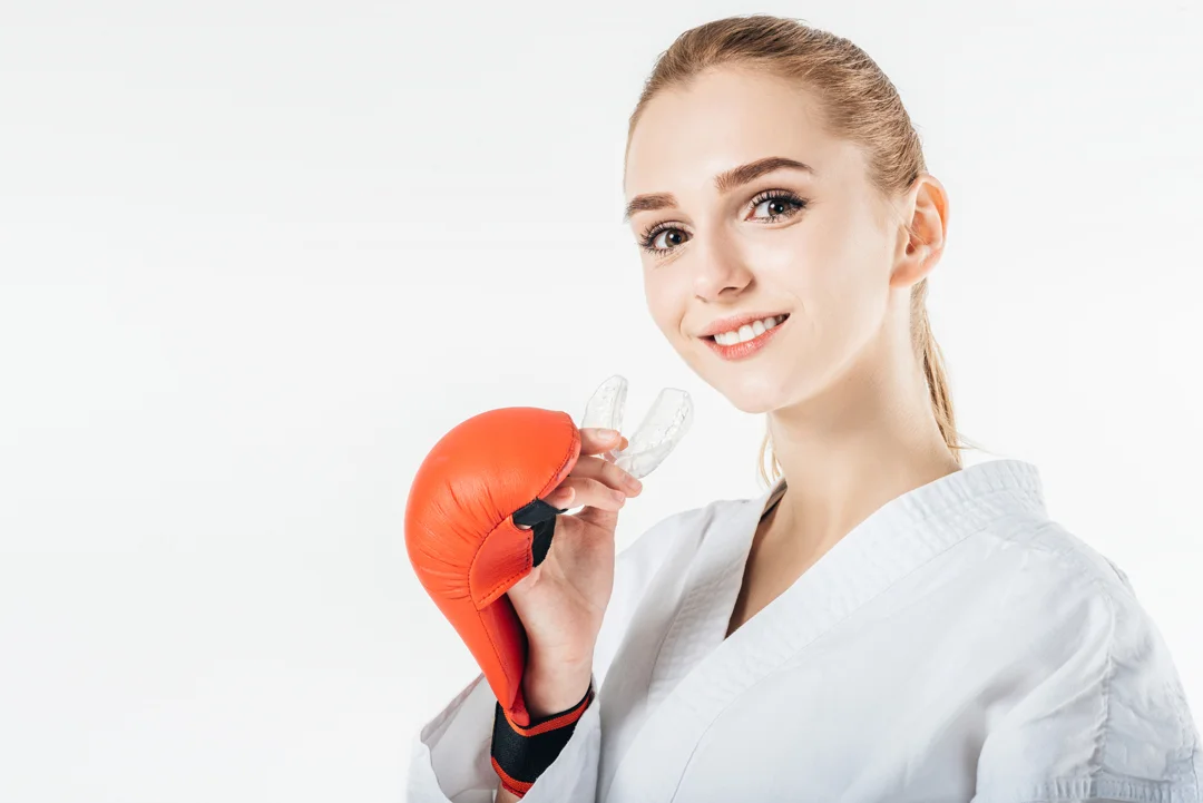 Smiling woman in a karate uniform holds a red boxing glove and a mouthguard against a white background.
