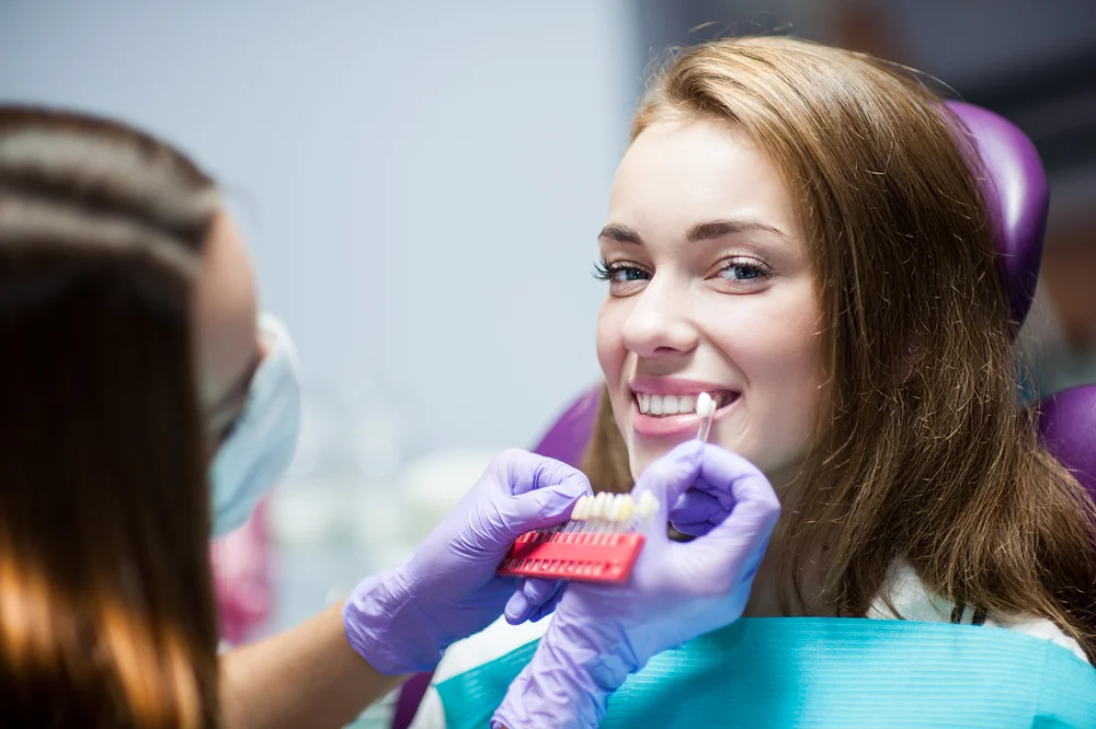 A dentist compares tooth shades to a smiling woman’s teeth during a dental appointment.