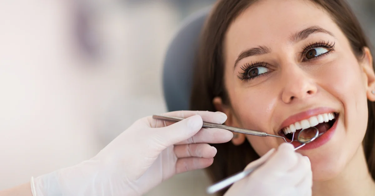 shutterstock_1052270732.jpg A woman smiles while a dentist examines her teeth with dental tools.