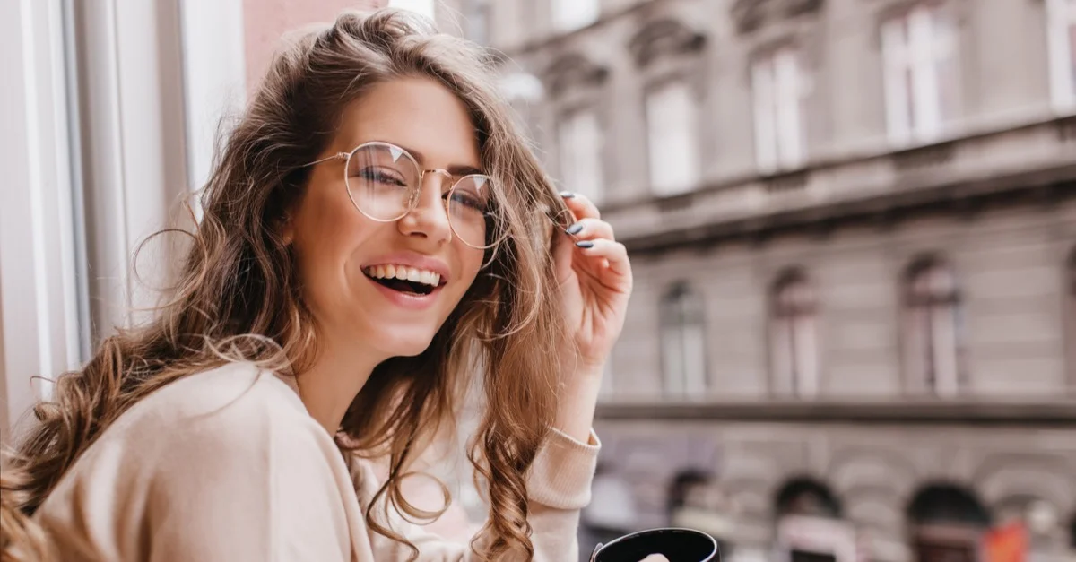 shutterstock_1082155934.jpg Smiling woman with long hair and glasses sits by a window, holding a coffee mug, in an urban setting.