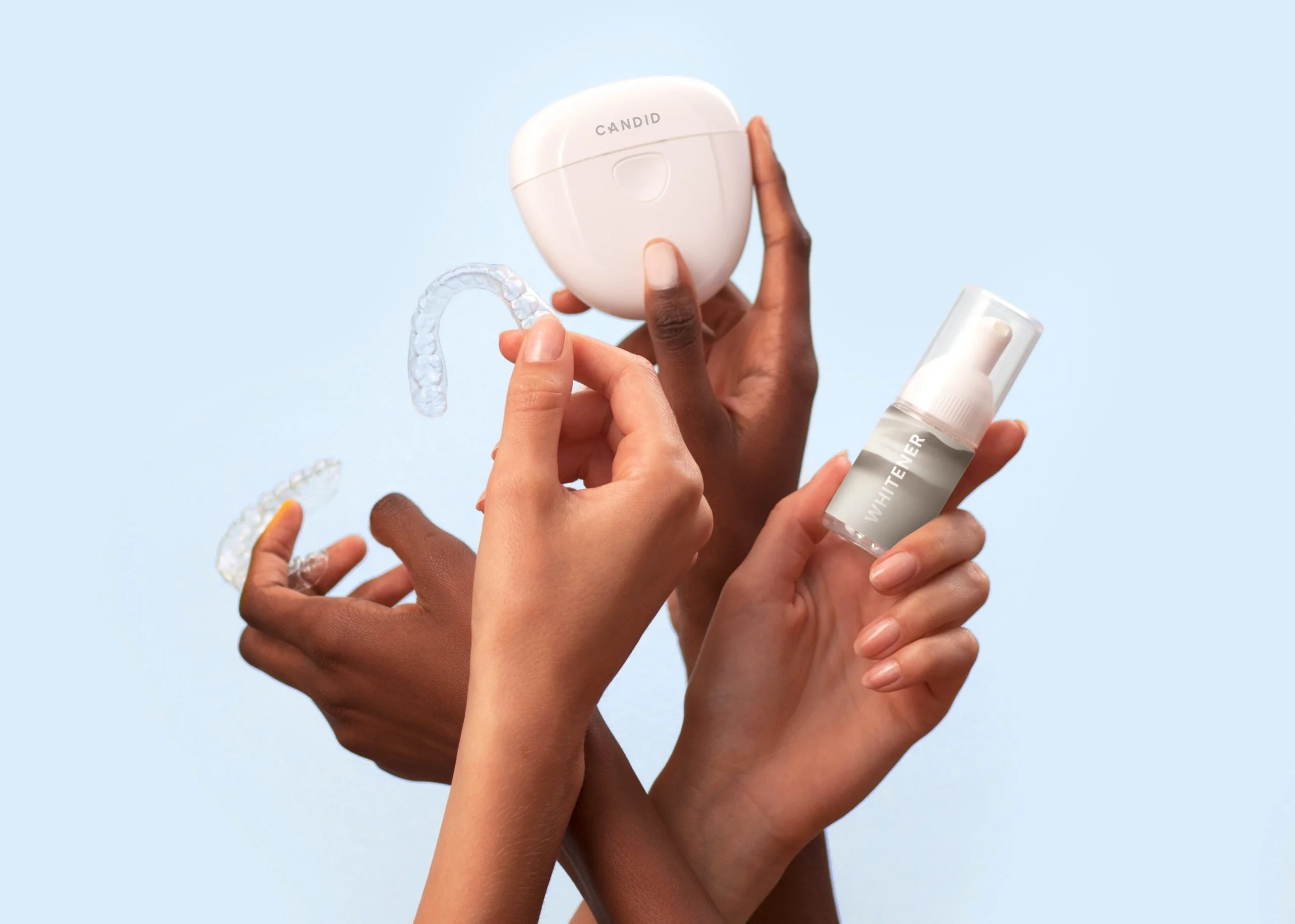 dental-retainer-scaled.jpg Four hands hold clear aligners, a Candid case, and a skincare bottle against a light blue background.