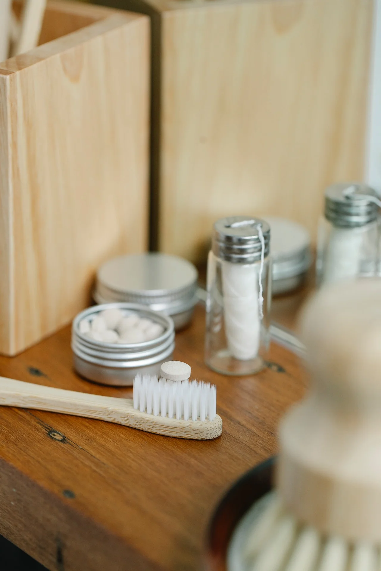 A bamboo toothbrush, toothpaste tablet, floss, and jars on a wooden bathroom counter near a mirror.