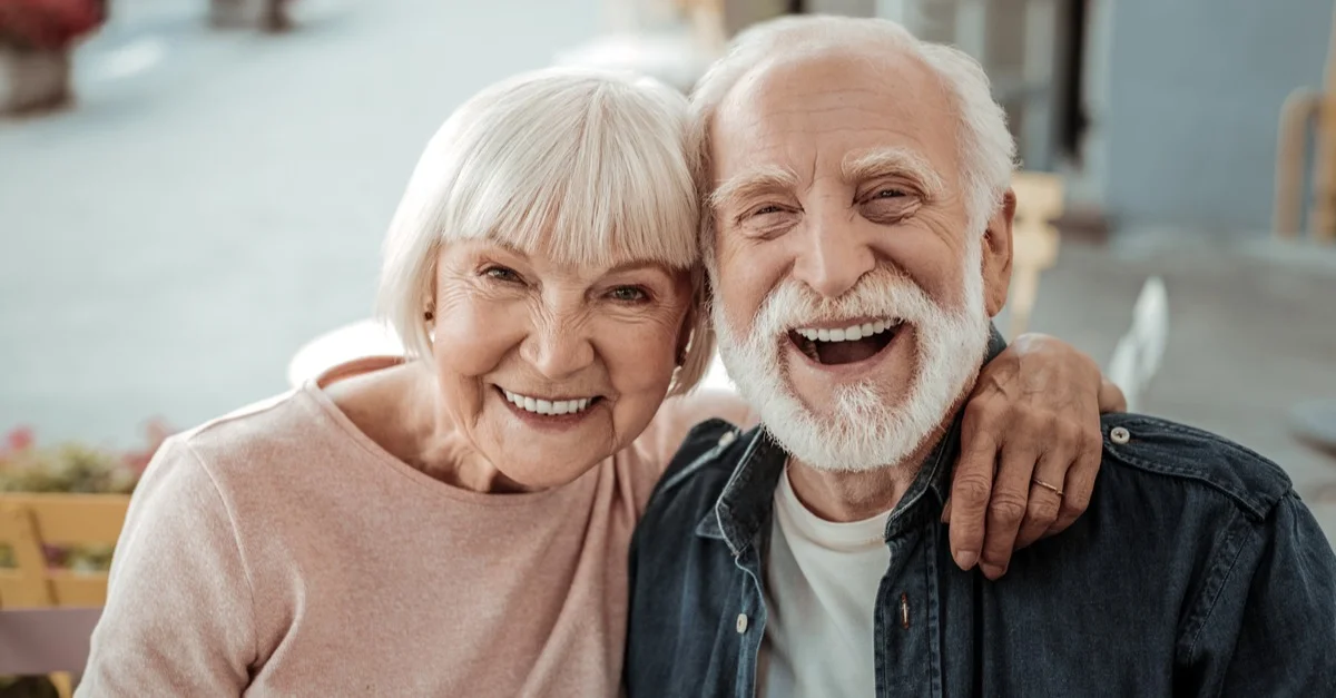 shutterstock_1565185228.jpg Smiling elderly couple sitting close together, with the woman's arm around the man's shoulders.