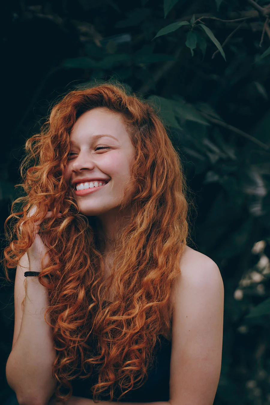 Woman with long curly red hair smiling, standing outdoors in front of dark green foliage.