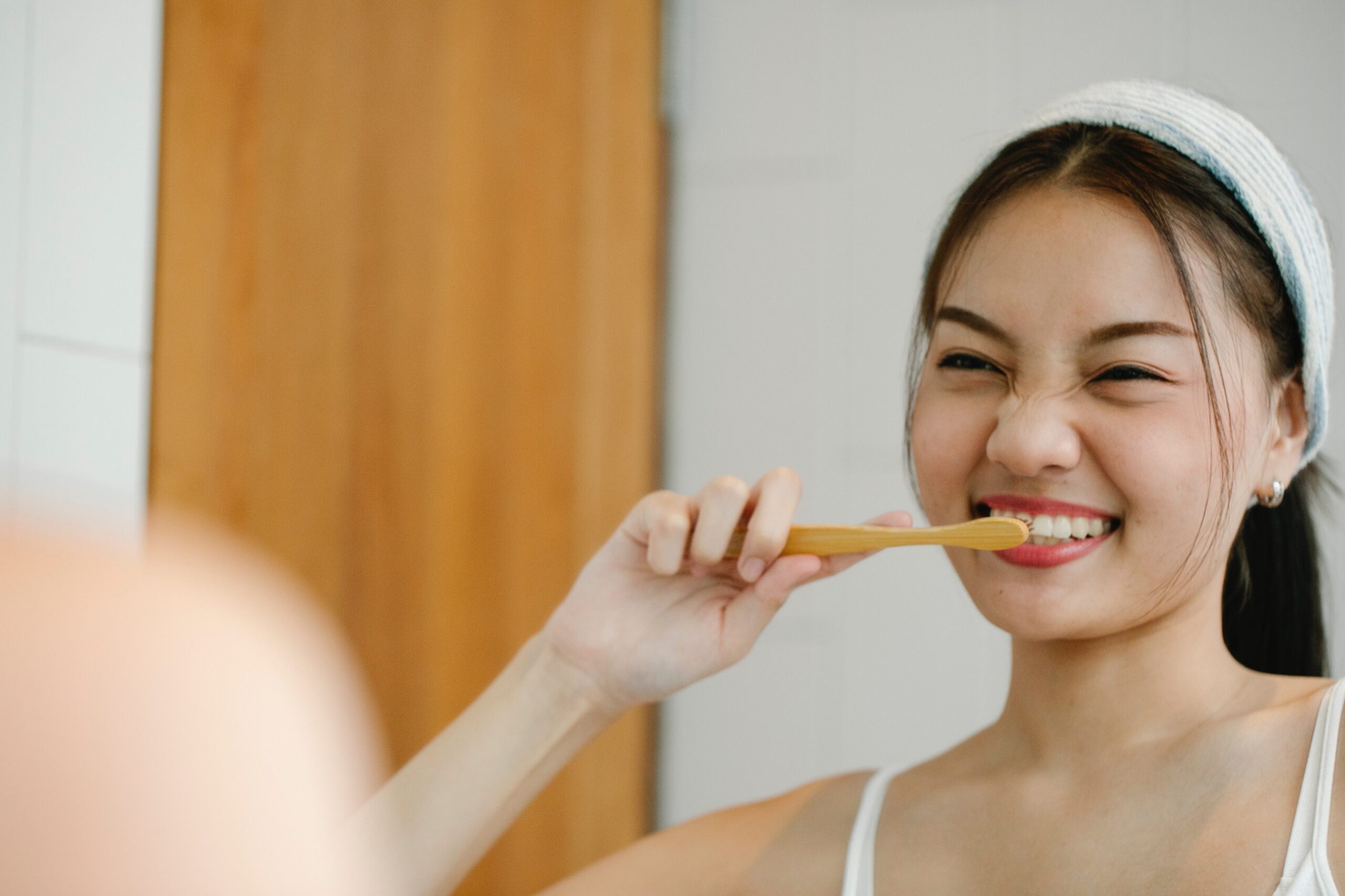 Woman smiling while brushing her teeth with a bamboo toothbrush in a bathroom.