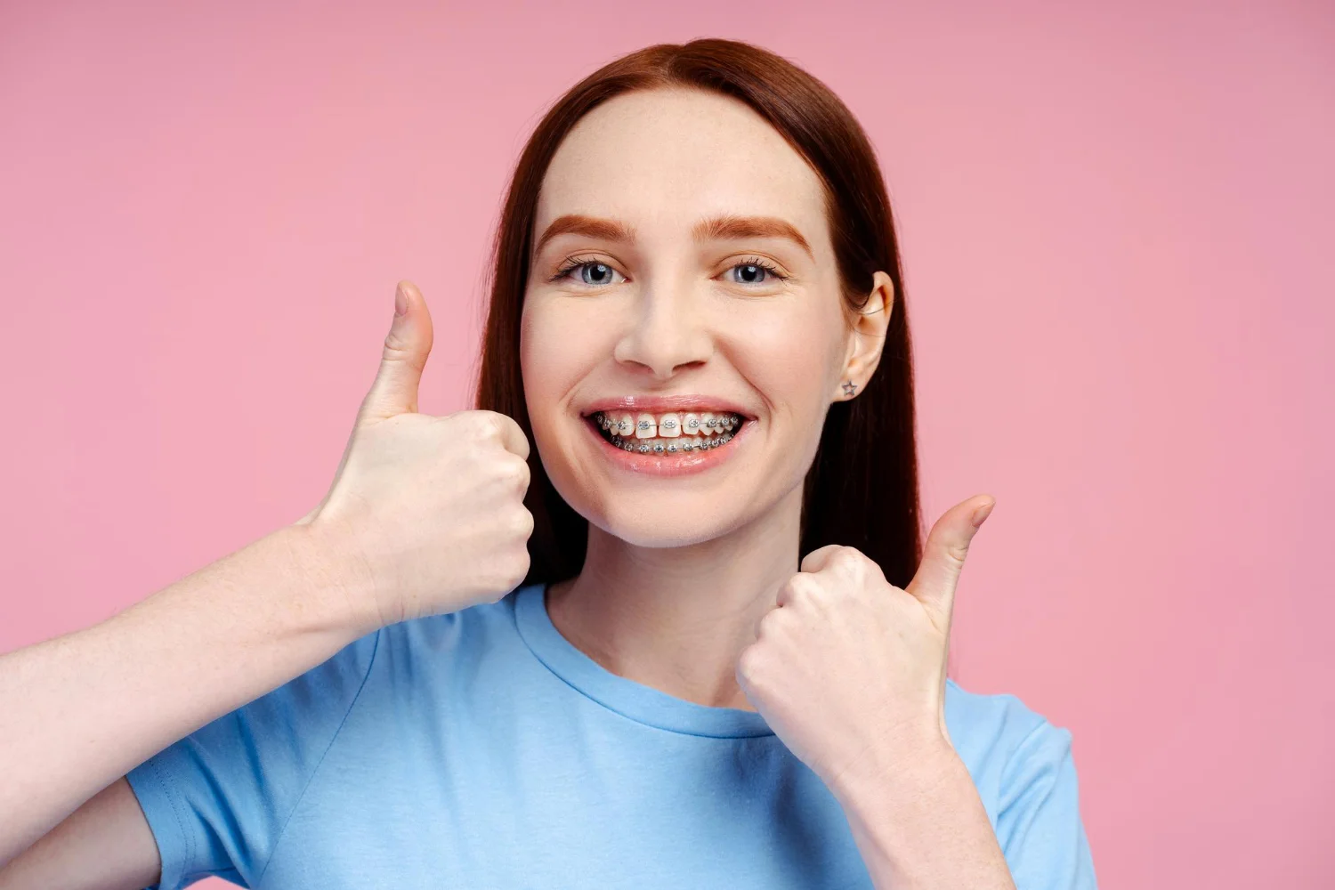 Smiling woman with braces in a blue shirt giving two thumbs up against a pink background.