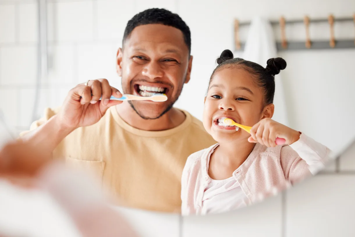 A man and a young girl smiling while brushing their teeth together in front of a bathroom mirror.