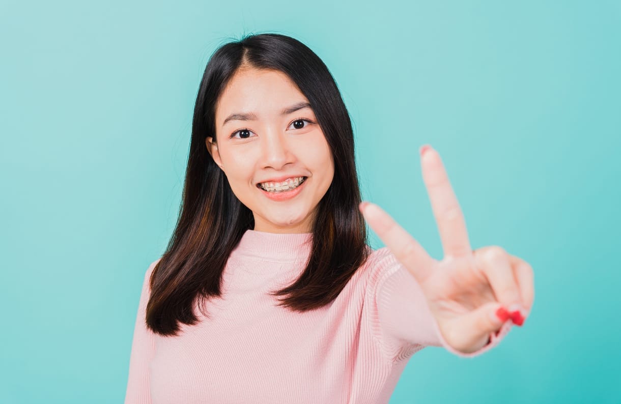 Invisalign vs. Braces Smiling woman with long dark hair and braces giving a peace sign on a blue background.