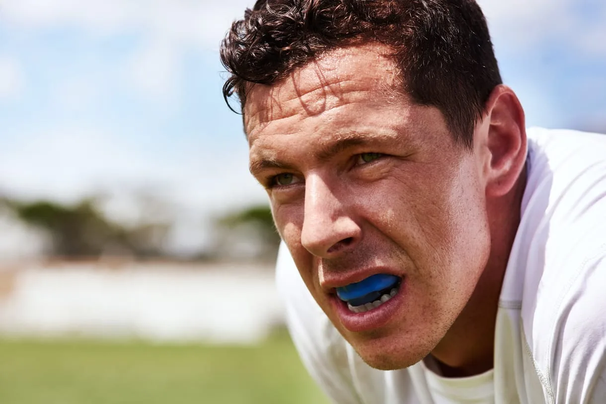 A man with a blue mouthguard rests outdoors, wearing a white shirt and looking determined.