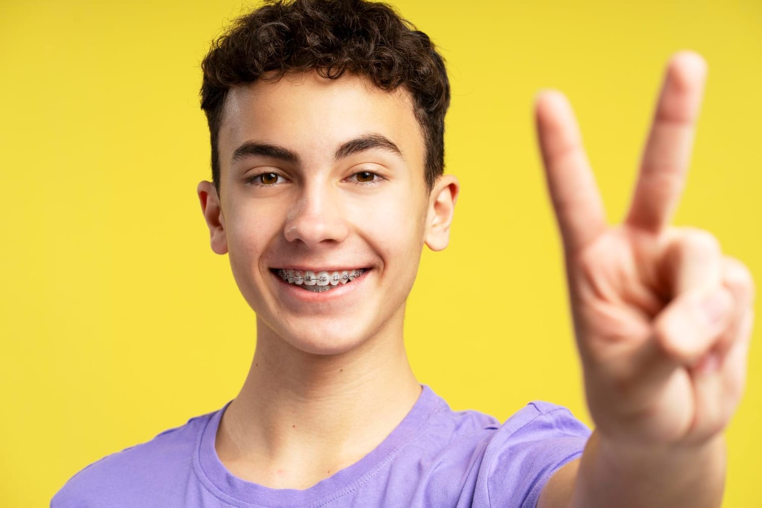 Teen boy with braces smiling, wearing a purple shirt, gives a peace sign with his fingers against a yellow background.