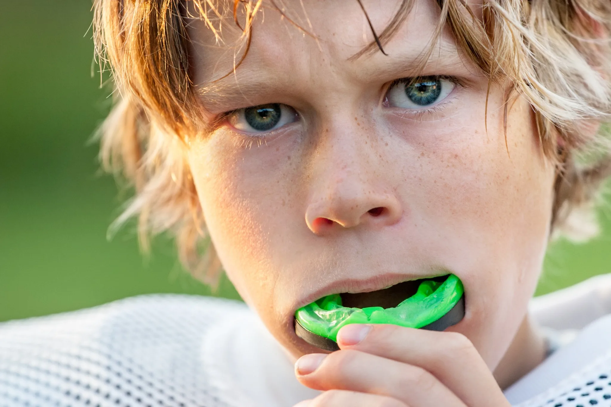 Close-up of a young boy in sports gear putting a green mouthguard in his mouth, looking serious.