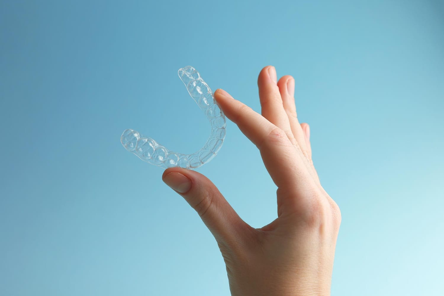 A hand holding a clear dental aligner, one of the types of dental retainers, against a blue background.