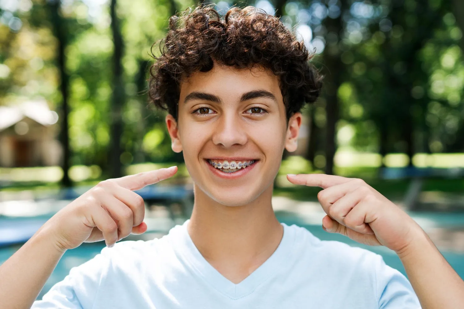 16182.jpg Teen boy with curly hair smiles and points to his braces outdoors, with trees blurred in the background.