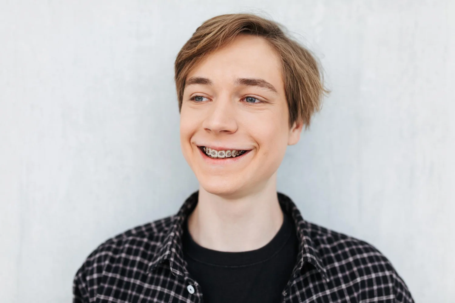 Smiling teenage boy with braces, wearing a black checkered shirt, standing against a light background.