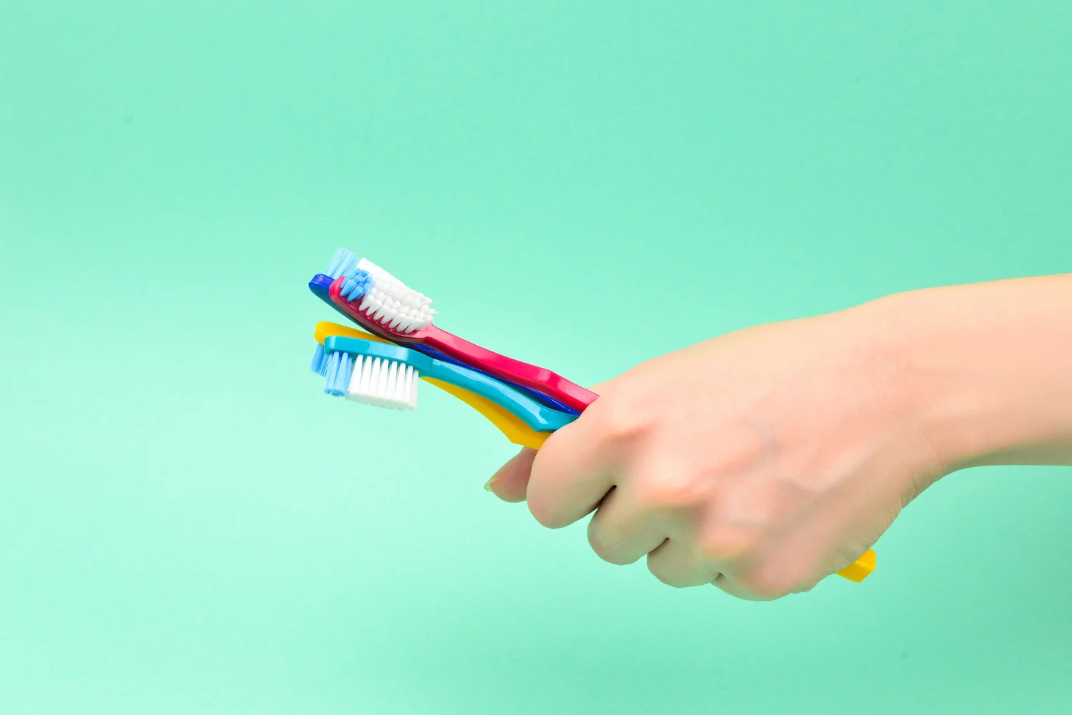 A hand holding three colorful toothbrushes against a light turquoise background.