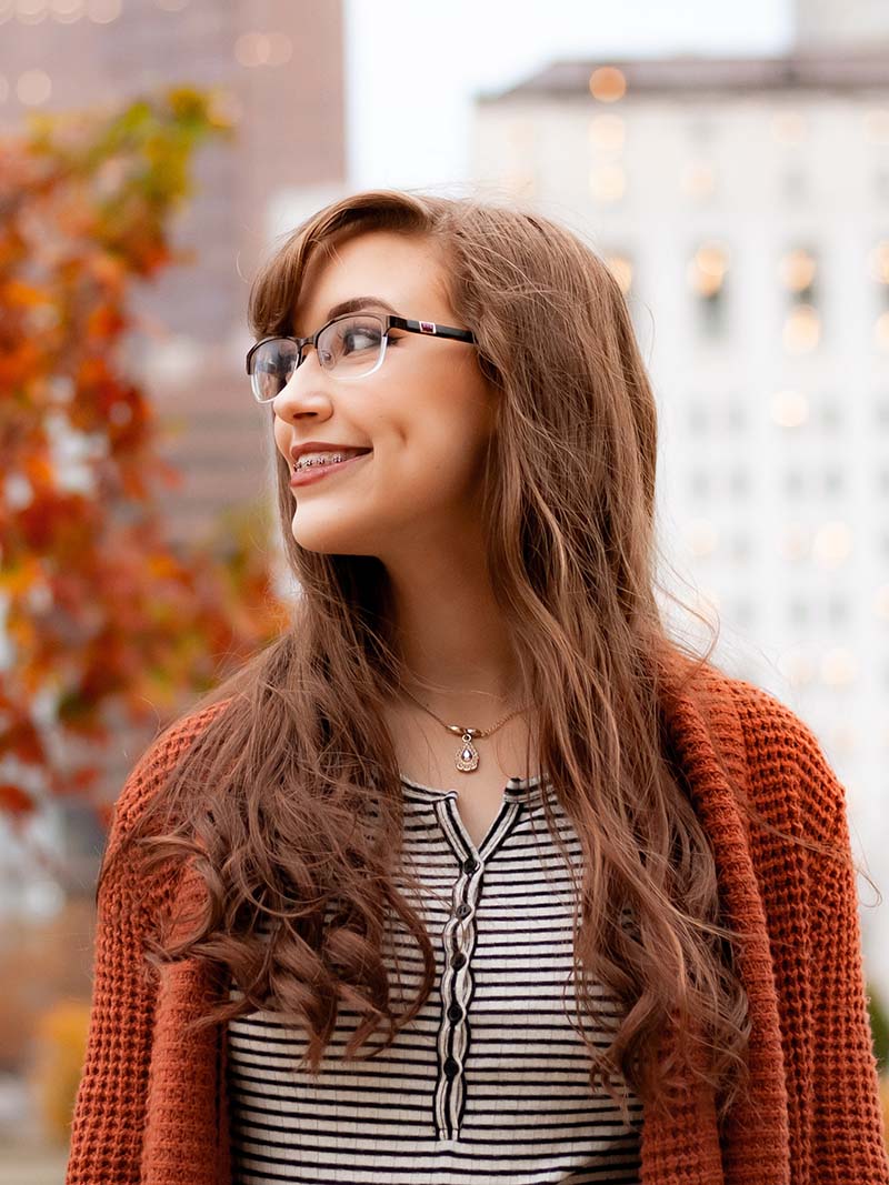 Smiling young woman with glasses and braces, wearing a striped shirt and orange sweater, outdoors in autumn.