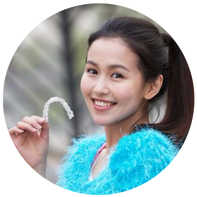 Woman smiling and holding a clear dental aligner, wearing a blue fuzzy top, outdoors.