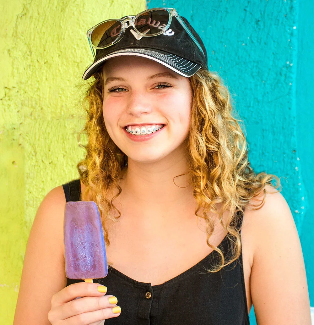 Smiling girl with braces holds a purple popsicle, wearing a hat and sunglasses, in front of a colorful wall.
