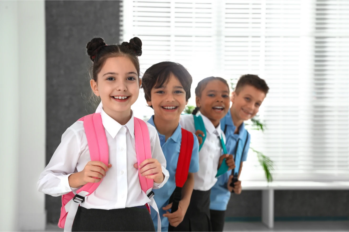 Four smiling schoolchildren with backpacks stand in a line indoors, ready for school.