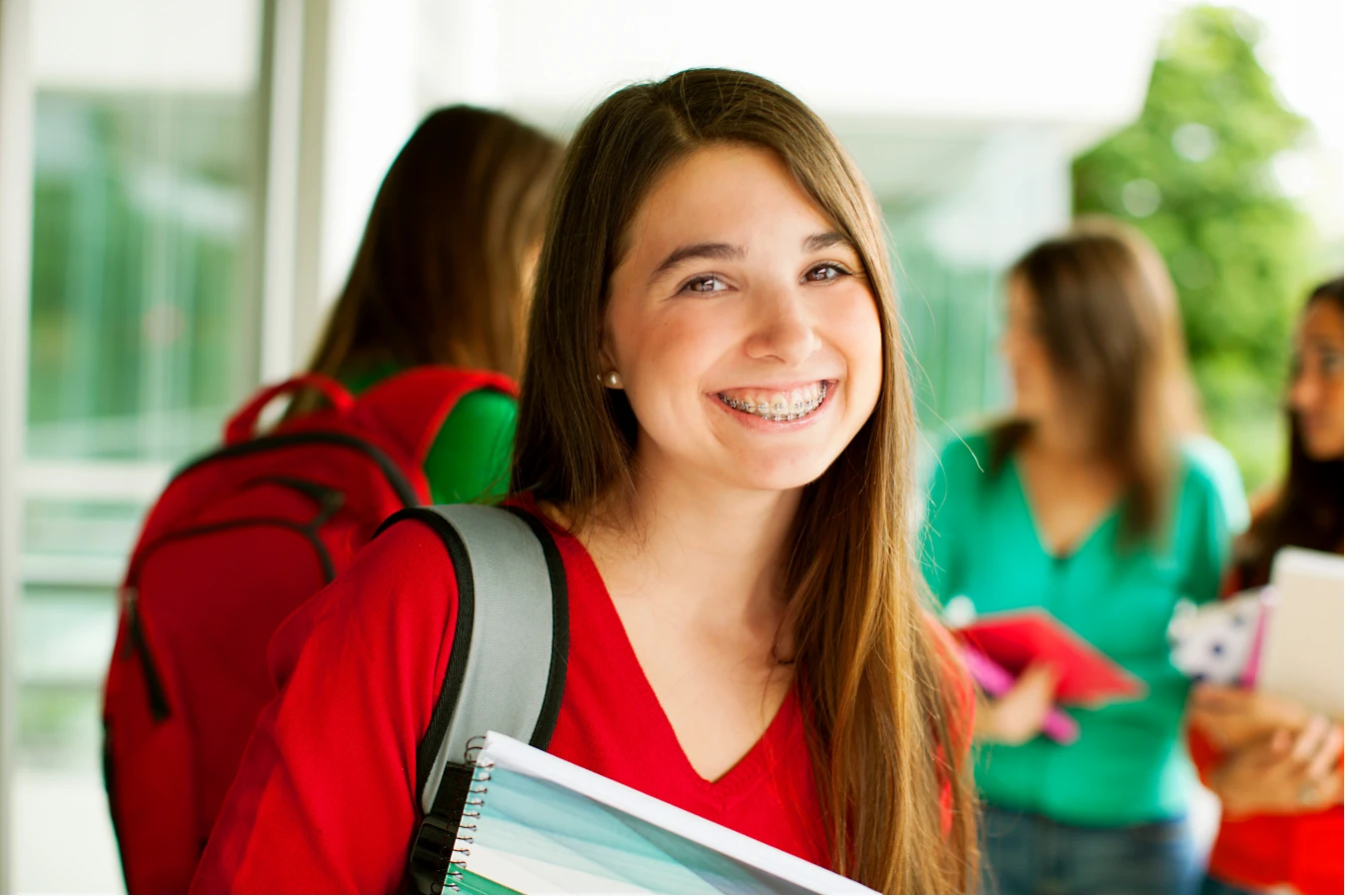 Smiling student with braces holding notebooks, wearing a red shirt and backpack, standing with friends.
