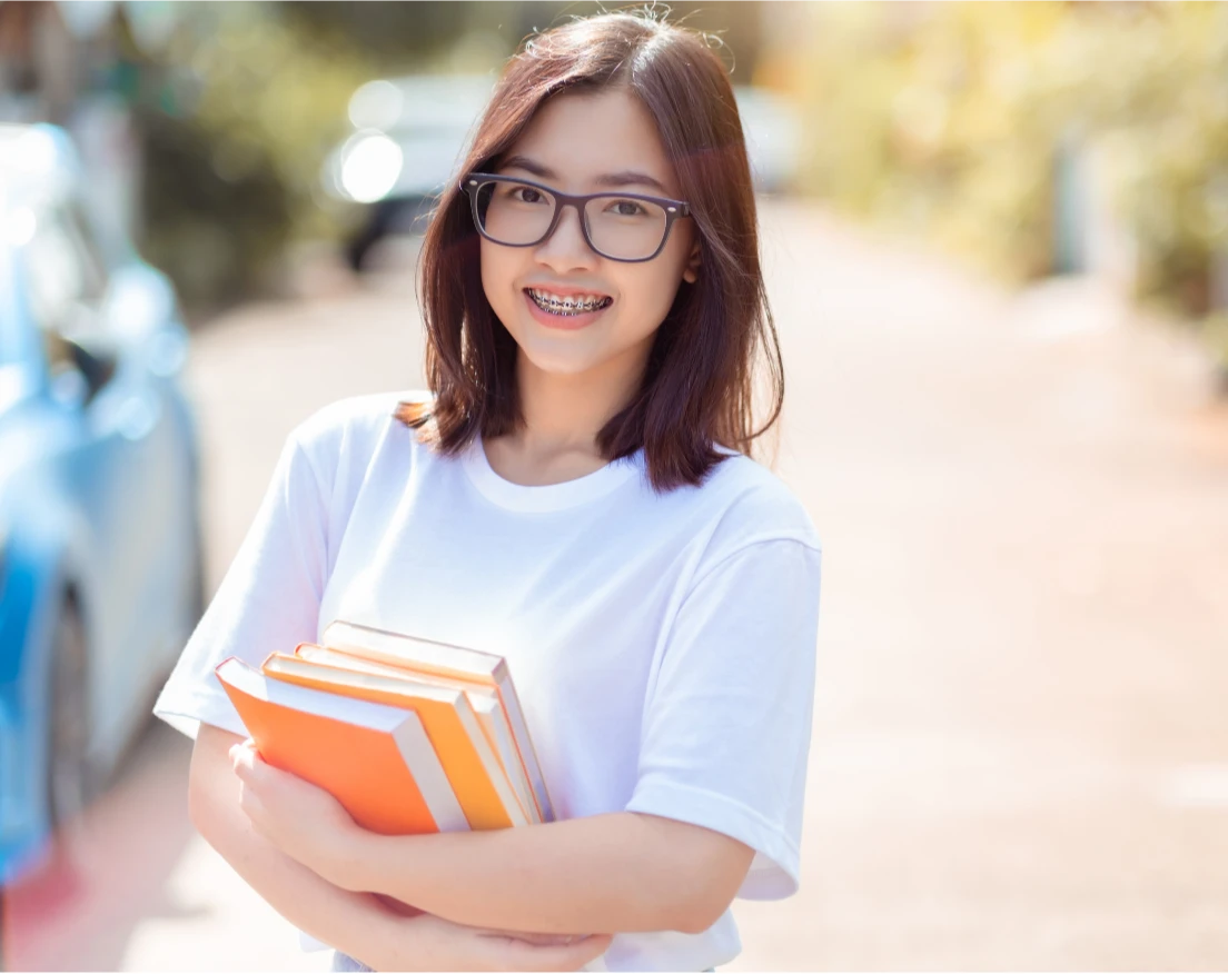Smiling young woman with glasses and braces holding books, standing outdoors on a sunny day.