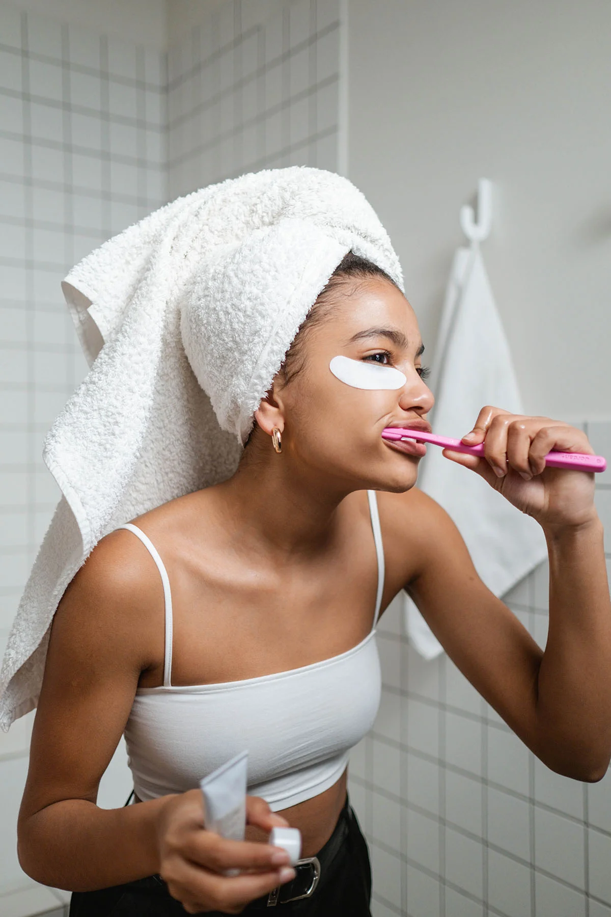 Woman with towel on head and eye patches brushes teeth in bathroom, wearing a white tank top.