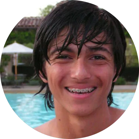 Smiling teenage boy with braces and wet hair standing in front of a swimming pool outdoors.