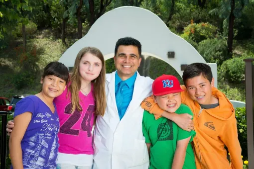 A man in a white coat stands smiling outdoors with four smiling children on a sunny day.