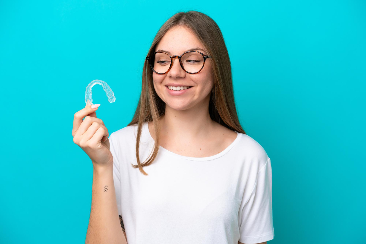 A woman wearing glasses looks at a clear plastic dental aligner in her hand.